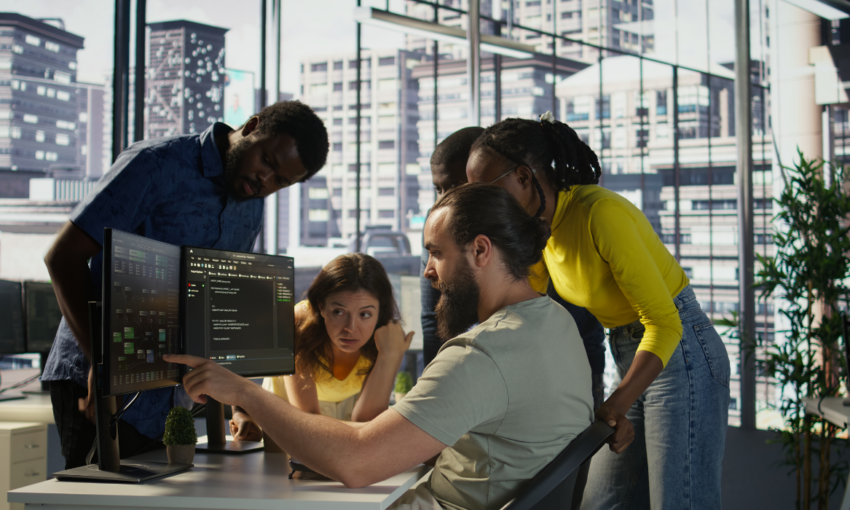 Diverse team of IT professionals collaborating on code at a computer in a modern office with city views.